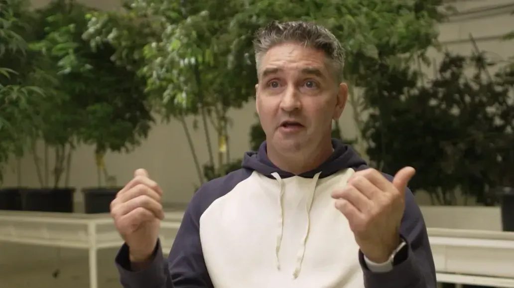 Man speaking with hands raised in front of indoor cannabis plants